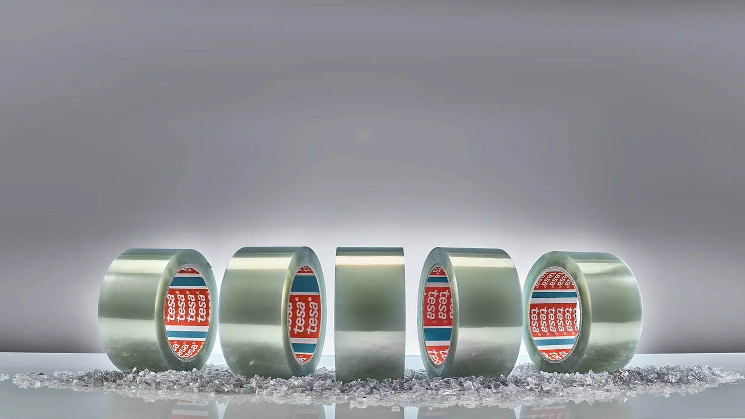 Five rolls of clear adhesive tesa tape are displayed vertically against a gray background. Small, scattered transparent granules are on the surface in front of the rolls. The tape cores are branded with red and blue labels. (This text has been generated by AI)