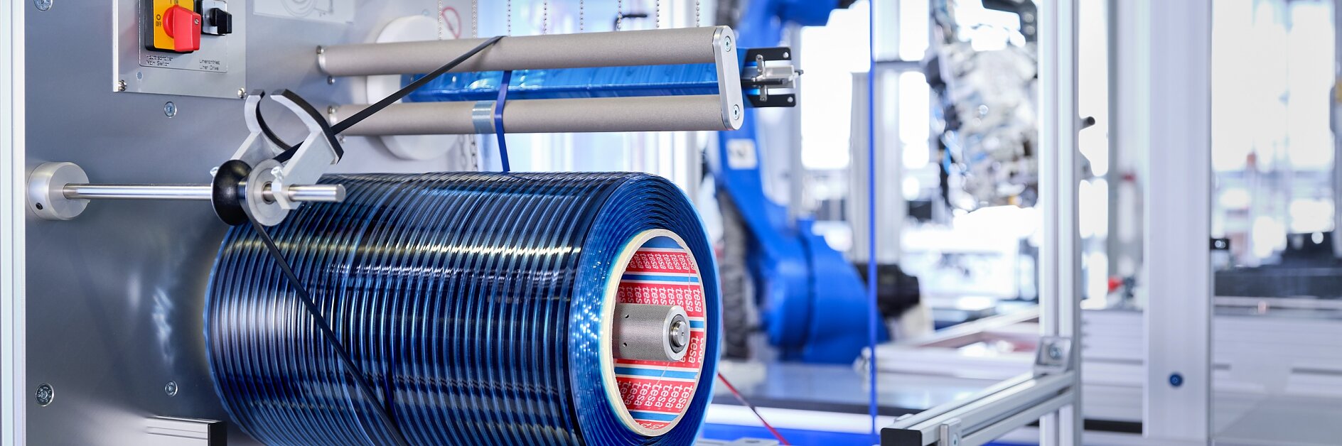 A manufacturing facility with industrial equipment, featuring a blue cylindrical component mounted on a metal frame using tesa tape. The machinery includes buttons, wires, and a robotic arm in the background. The setting is clean and organized.
