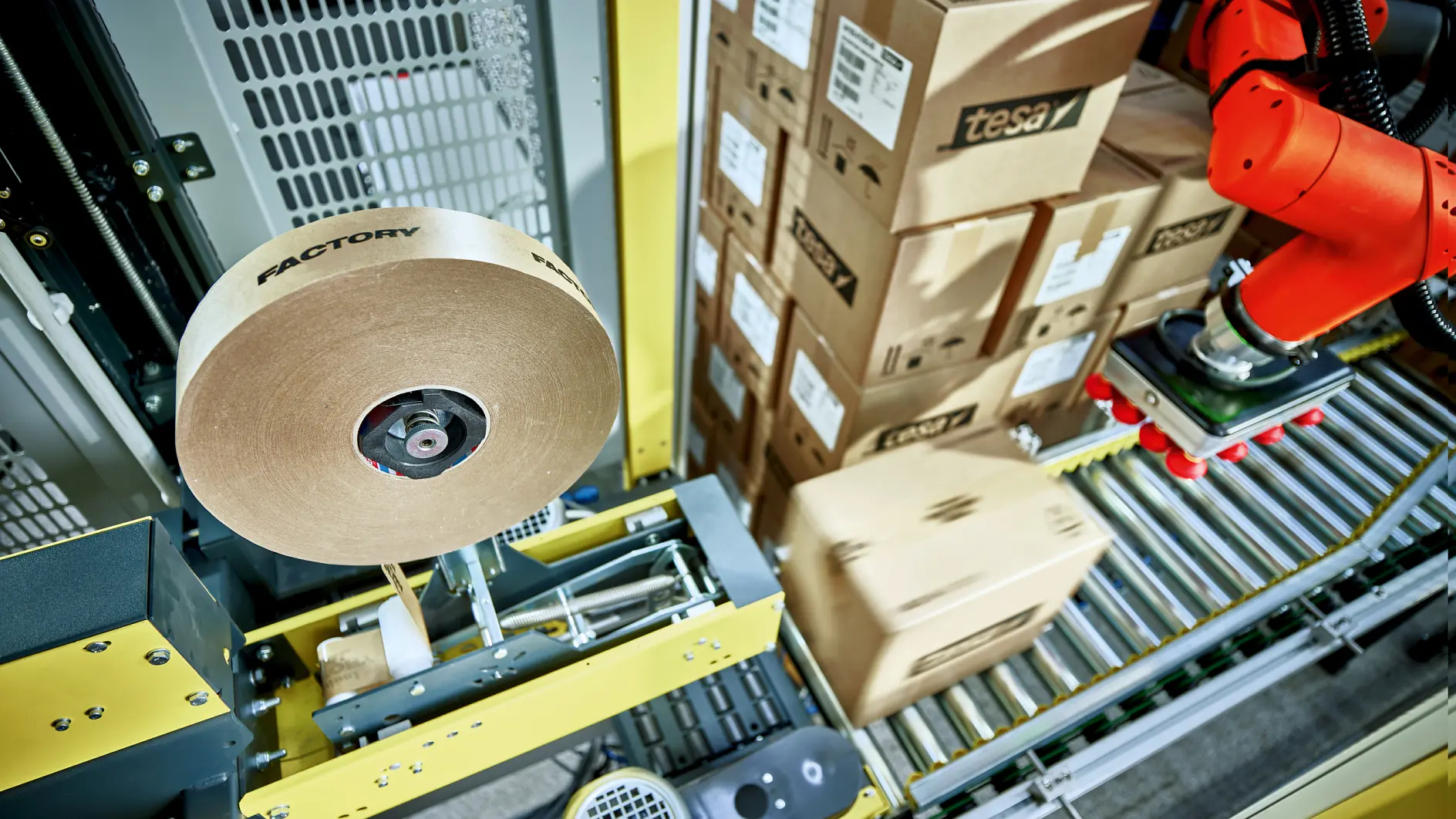 A robotic arm is operating in a factory setting, handling cardboard boxes on a conveyor system. A roll of tesa tape labeled factory is part of the machinery. Stacked boxes in the background are marked with tesa.