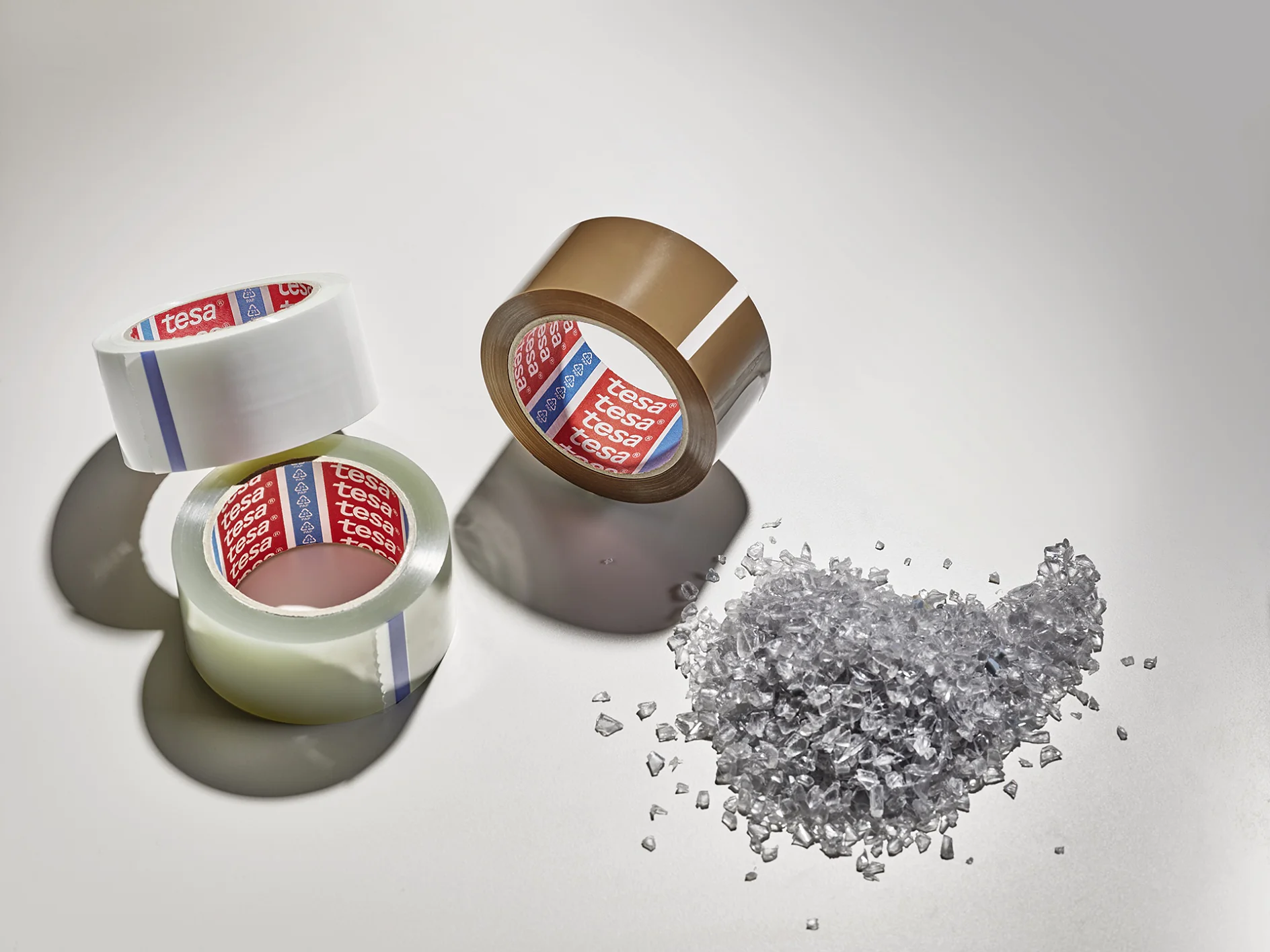 Three rolls of packing tesa tape, two clear and one brown, are arranged on a light surface. Beside them is a small pile of clear plastic granules. Shadows extend from the objects. (This text has been generated by AI)