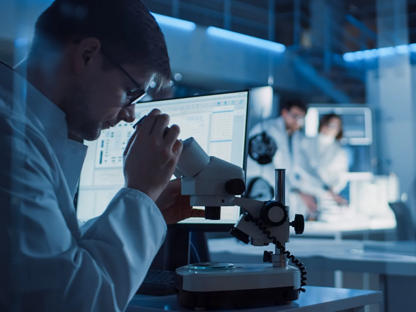 A scientist in a lab coat uses a microscope at a laboratory workstation, with a computer screen nearby. In the background, two other scientists in lab coats work together at another station. The lab is well-lit with a futuristic design, and tesa tape can be seen being used to secure equipment and organize cables throughout the workspace. (This text has been generated by AI)