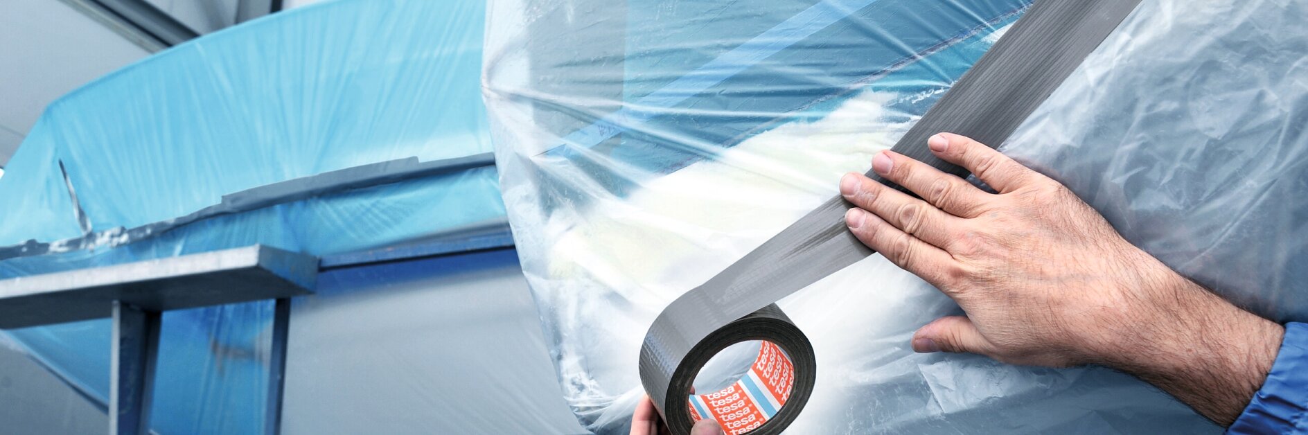 Two hands apply gray tesa tape to secure clear plastic sheeting over a large blue object, possibly in an industrial or construction setting. The background shows metal scaffolding and parts of the structure.