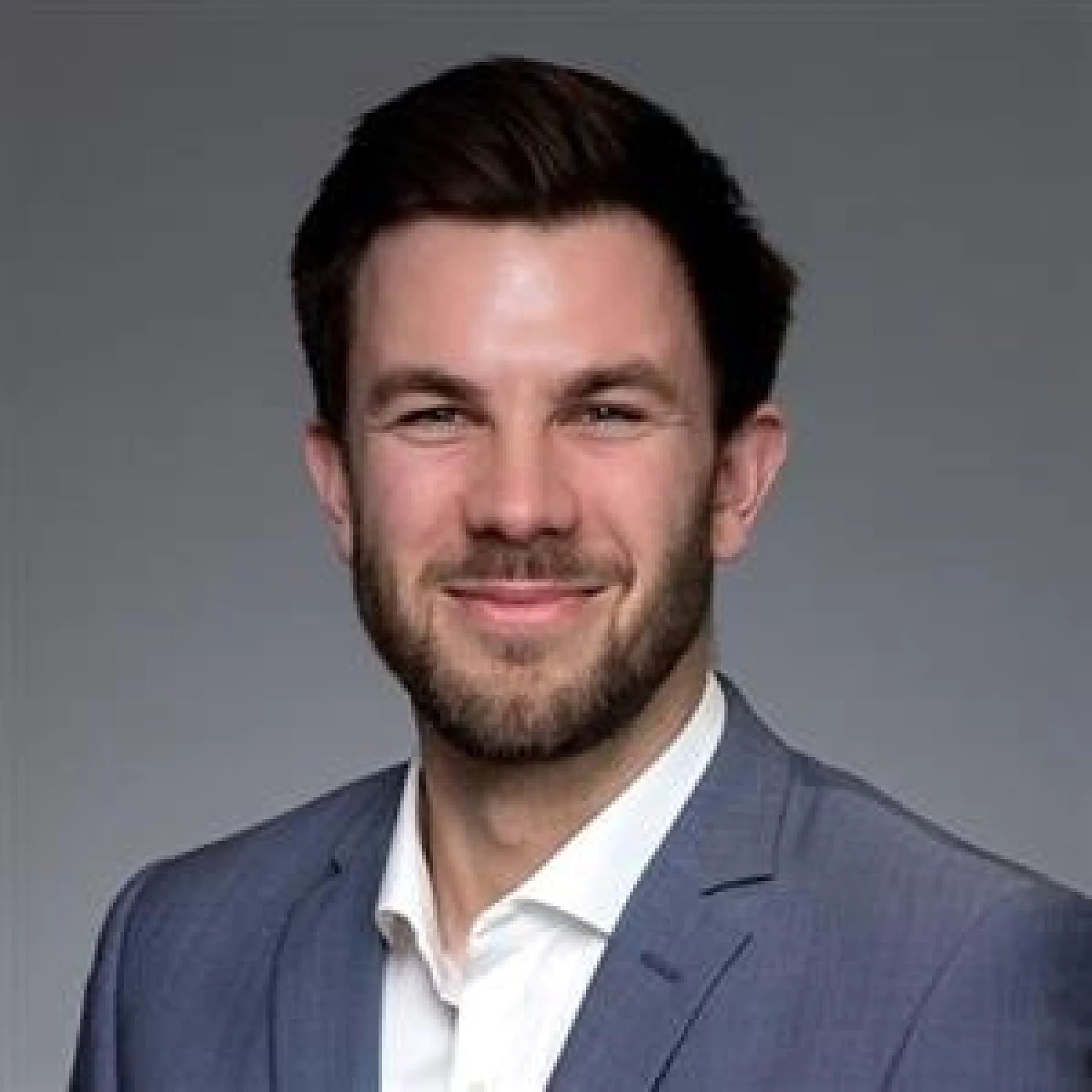 Man with short dark hair and beard wearing a light blue suit jacket and white shirt, smiling at the camera against a plain gray background. (This text has been generated by AI)