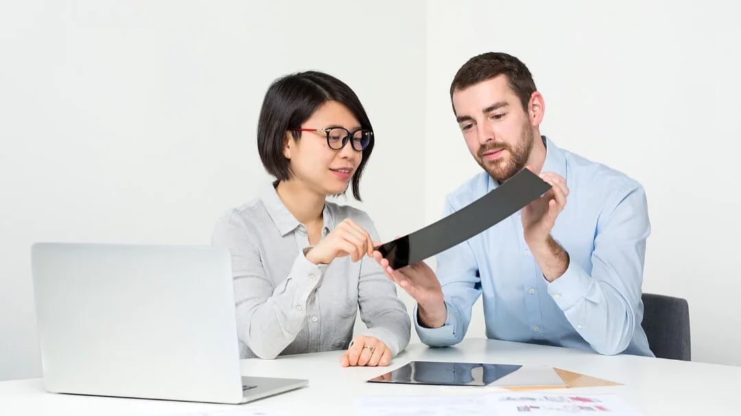 Two people at a table examining a flexible screen. The woman points to the screen while the man holds it. A laptop and paper with diagrams are on the table, along with some tesa tape. Both individuals are focused on the screen, using tesa tape as part of their setup. (This text has been generated by AI)