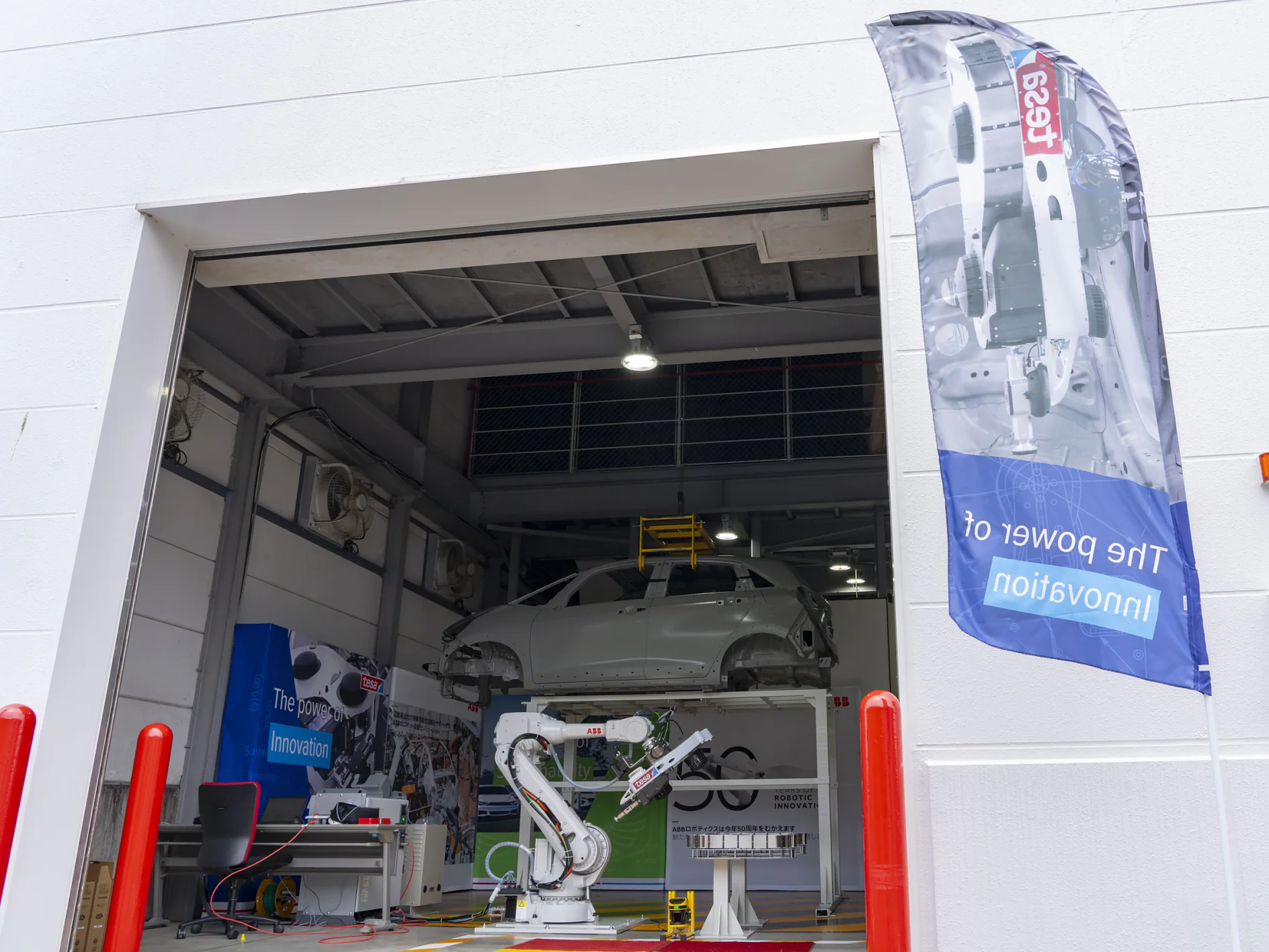 A car is elevated inside an open garage with a robotic arm visible in front. A banner outside displays a partially visible image and the text The Power of Innovation. The setting appears industrial, with red poles flanking the entrance. (This text has been generated by AI)
