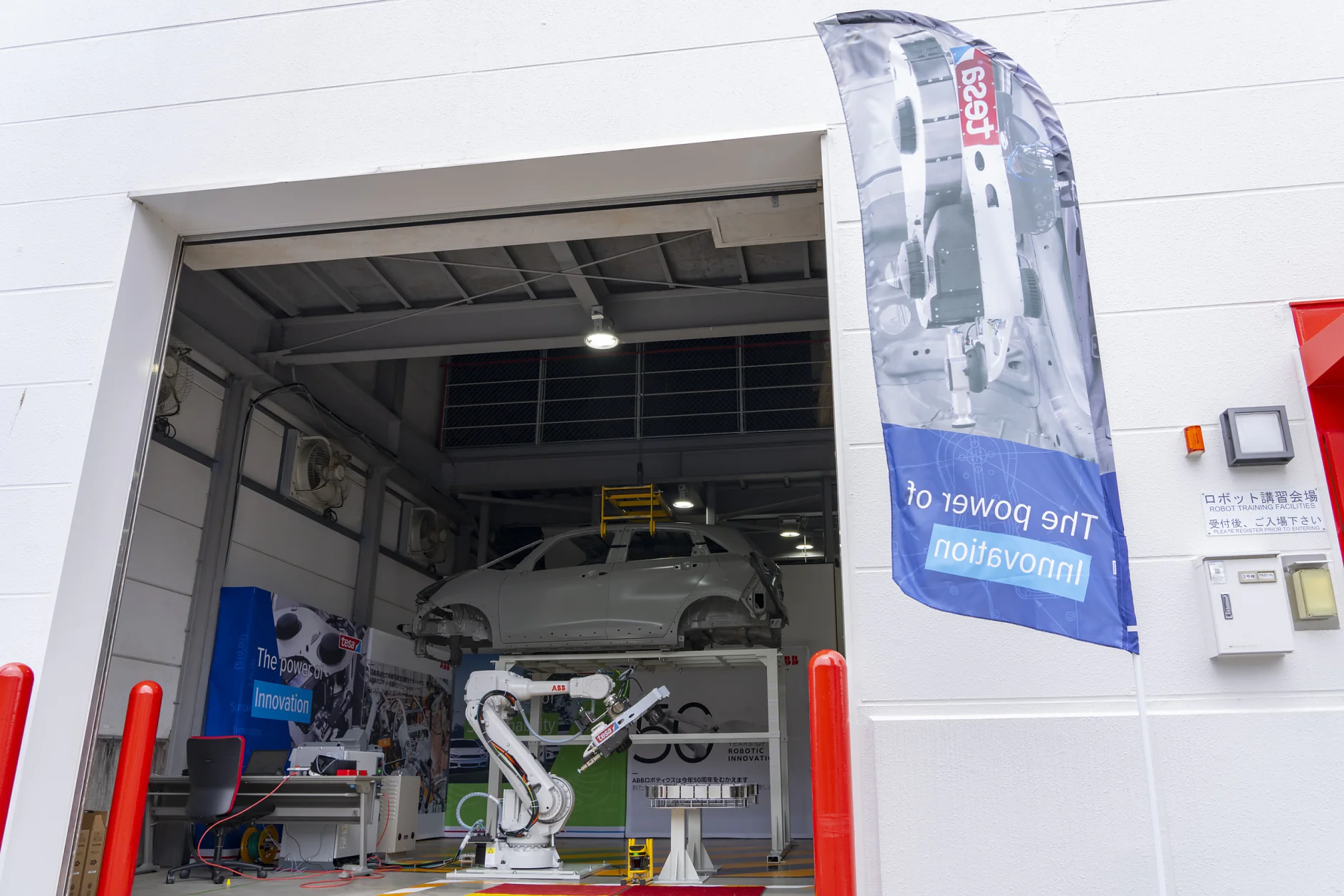 A car is elevated inside an open garage with a robotic arm visible in front. A banner outside displays a partially visible image and the text The Power of Innovation. The setting appears industrial, with red poles flanking the entrance. (This text has been generated by AI)