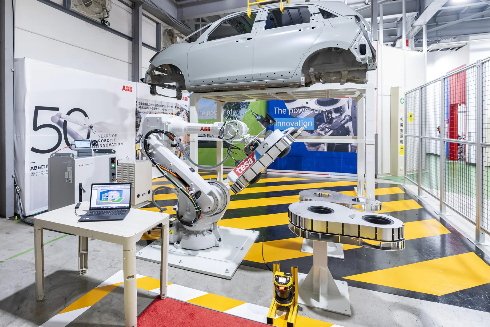 A robotic arm operates in an automotive assembly line with a car frame positioned above. A laptop is visible on a nearby table, while two circular machines and a banner displaying The power of innovation are in the background. (This text has been generated by AI)