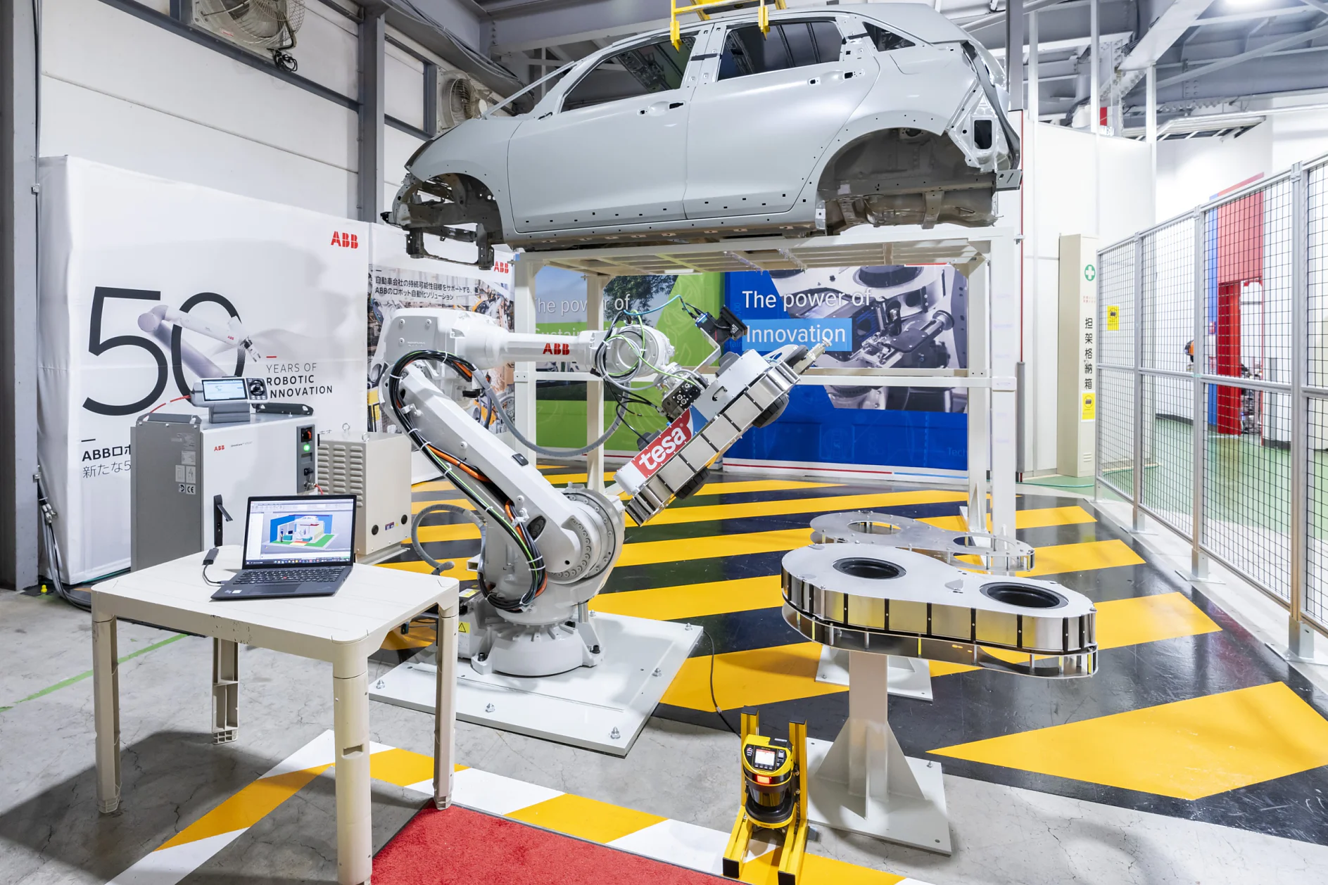 A robotic arm operates in an automotive assembly line with a car frame positioned above. A laptop is visible on a nearby table, while two circular machines and a banner displaying The power of innovation are in the background. (This text has been generated by AI)