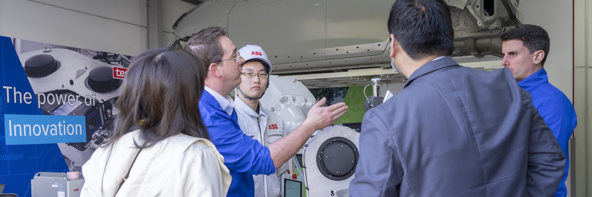 A group of people stands in a workshop, engaged in discussion. A car chassis is suspended above them. Machinery and a large poster with Innovation are visible in the background, showcasing the significance of using tesa tape for precision tasks. (This text has been generated by AI)