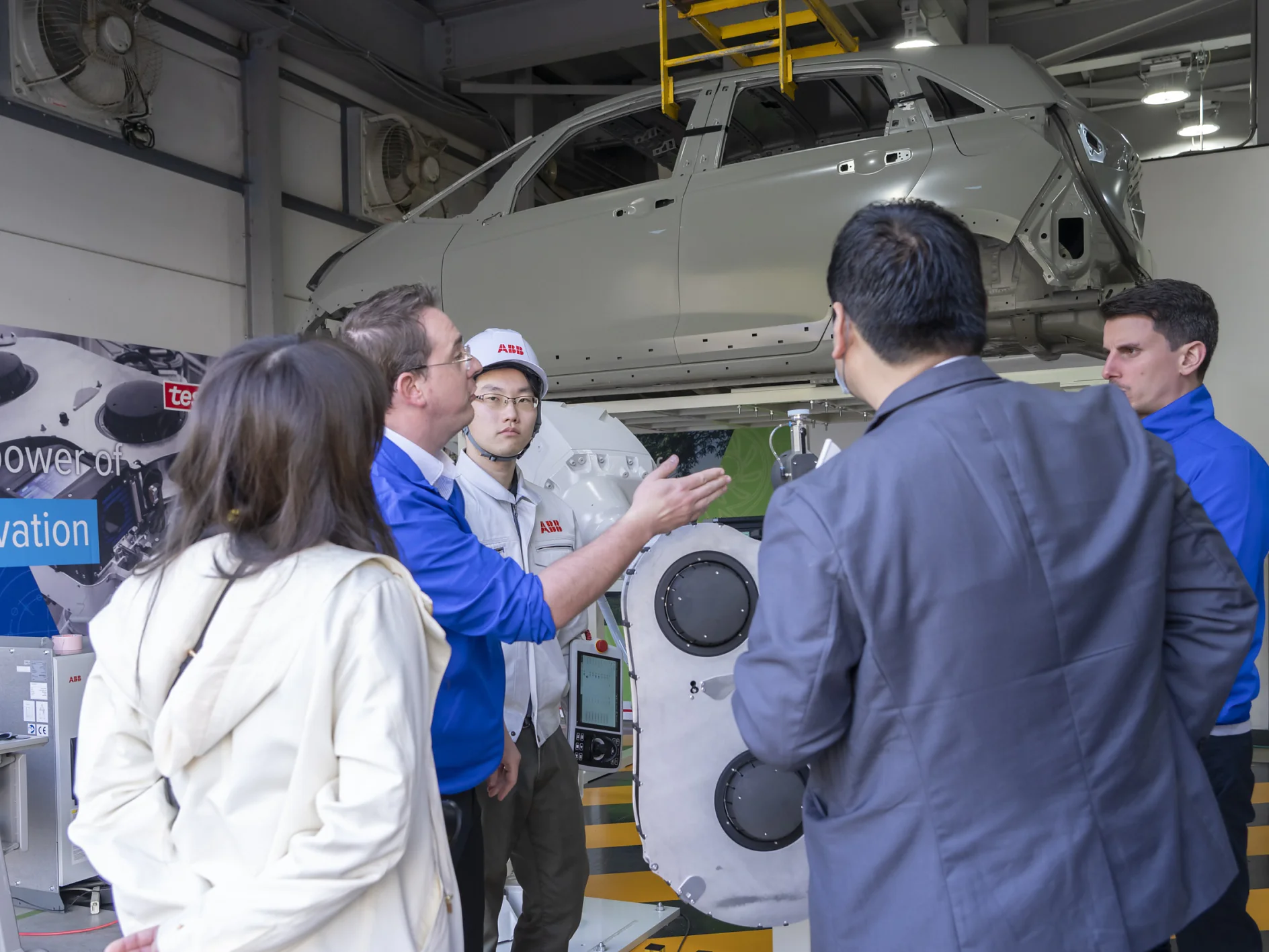 A group of people stands in a workshop, engaged in discussion. A car chassis is suspended above them. Machinery and a large poster with Innovation are visible in the background, showcasing the significance of using tesa tape for precision tasks. (This text has been generated by AI)