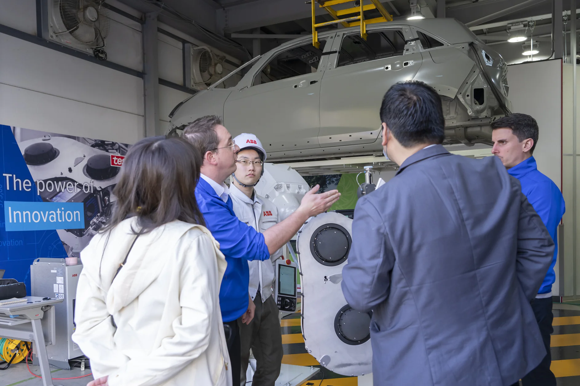 A group of people stands in a workshop, engaged in discussion. A car chassis is suspended above them. Machinery and a large poster with Innovation are visible in the background, showcasing the significance of using tesa tape for precision tasks. (This text has been generated by AI)