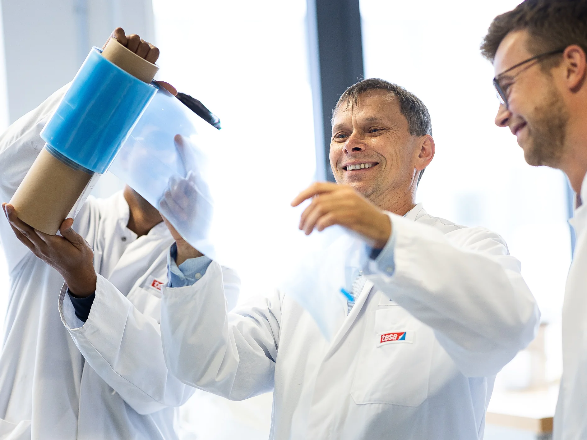 Three people in lab coats examine a roll of transparent blue tesa tape in a bright laboratory setting. One person holds the tesa tape upright, while another pulls a section away. They appear focused and engaged. (This text has been generated by AI)