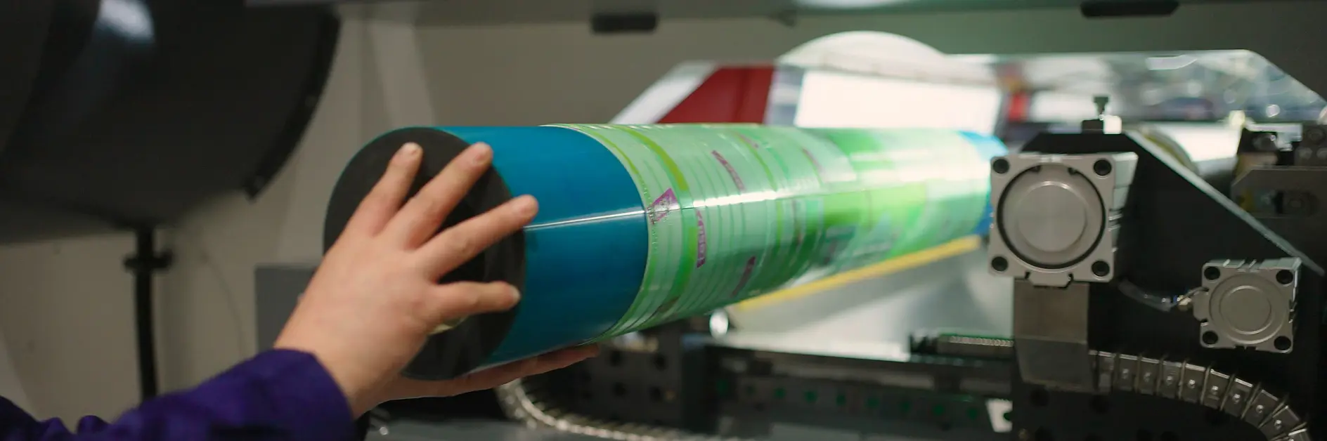 A person places a cylindrical green and blue printing plate into a large printing machine using tesa tape. The machine features various buttons and indicators. The setting seems to be in an industrial or manufacturing environment.