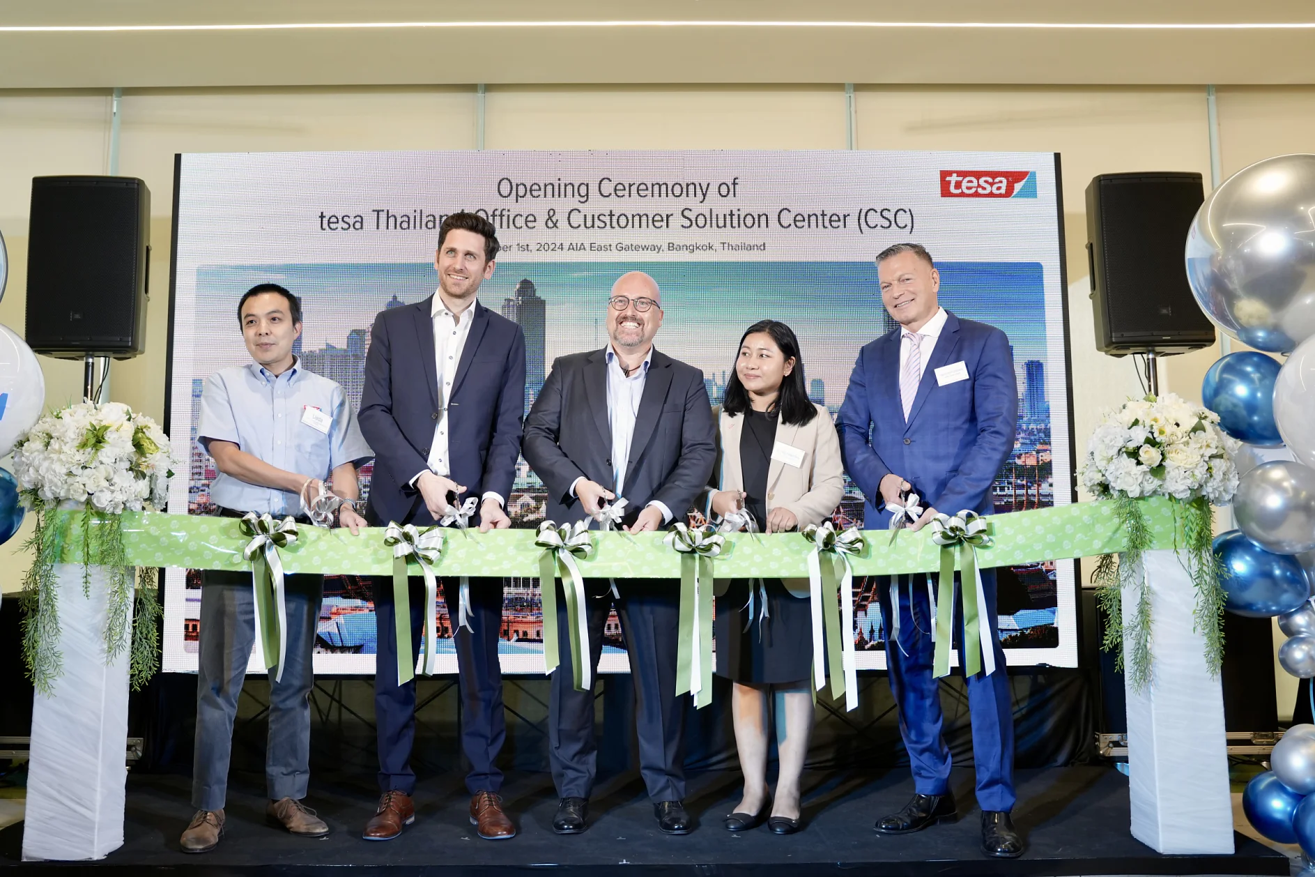 Traditional “Ribbon Cutting Ceremony” for the opening of the new spaces in Bangkok, Thailand Five people stand in front of a sign at the opening ceremony of the tesa Thailand Office & Customer Solution Center. They are cutting a ribbon with scissors. There are floral arrangements and balloons on either side of them. (This text has been generated by AI)