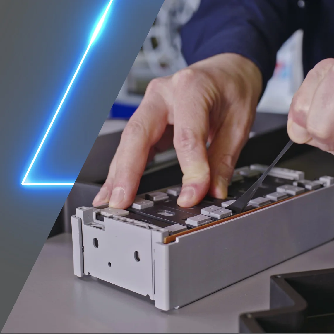 A person is handling a metallic component on a workbench, using a tool for precision. The component has a rectangular shape with several slots and raised areas. A blue, diagonal neon line overlays the image on the left side, highlighting the use of tesa tape in securing elements during the process. (This text has been generated by AI)