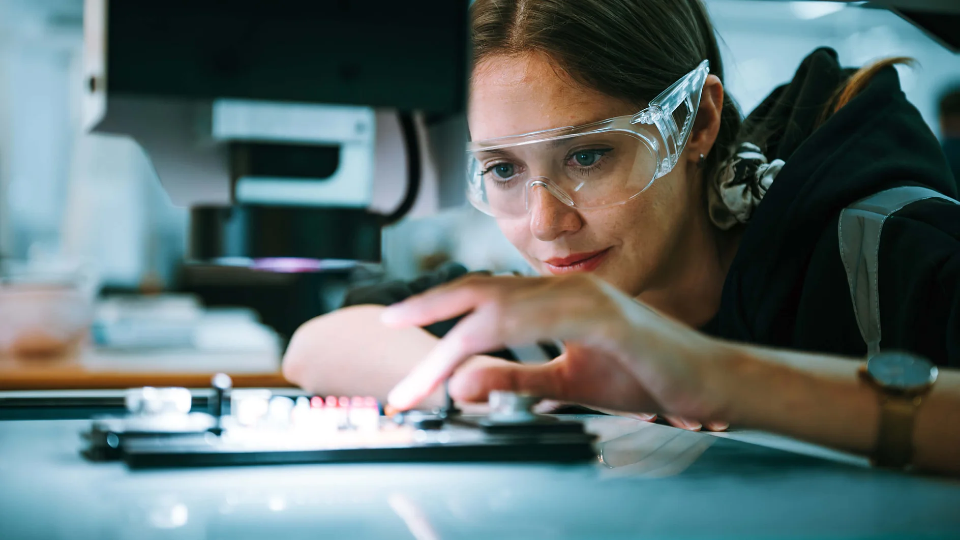 ARVR Foldable LP_High Performance A woman wearing safety goggles is intently working with a piece of machinery. Her hand is adjusting a small component on a device using tesa tape, and she is focused on the task. The background is blurred, suggesting an industrial or laboratory setting. (This text has been generated by AI)