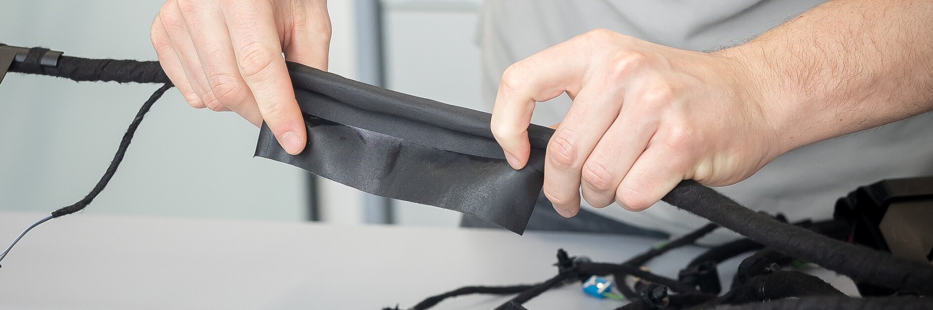 A person in a white shirt is wrapping black electrical tesa tape around a bundle of wires or cables on a table. Their hands are pulling the tesa tape tightly to secure the cables. (This text has been generated by AI)