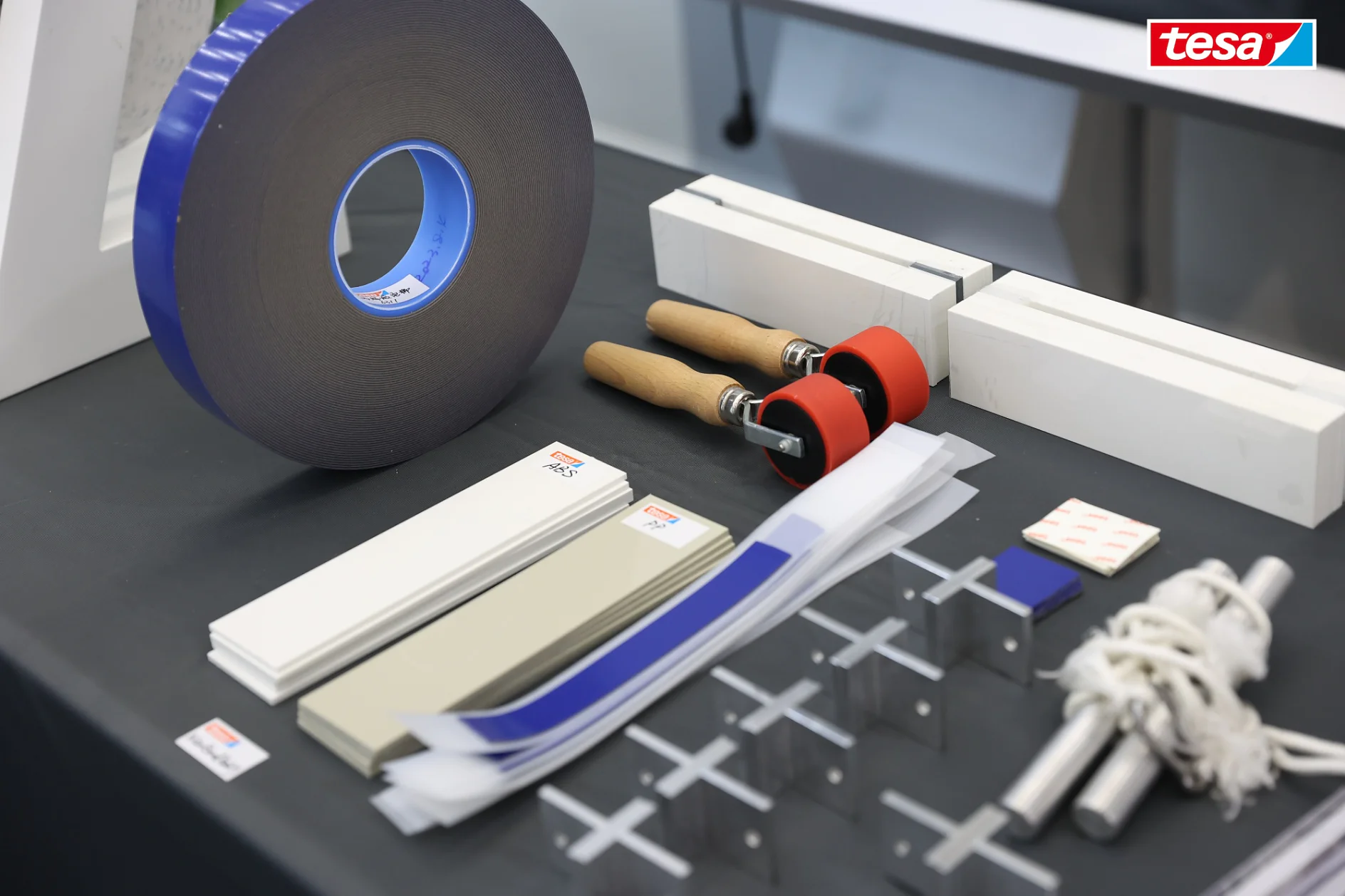 A table displays tesa tape rolls, utility knives, white strips, small labeled cards, plastic strips, metal rods, cross-shaped acrylic pieces, rope, and a product sample booklet, with a tesa logo in the corner. (This text has been generated by AI)