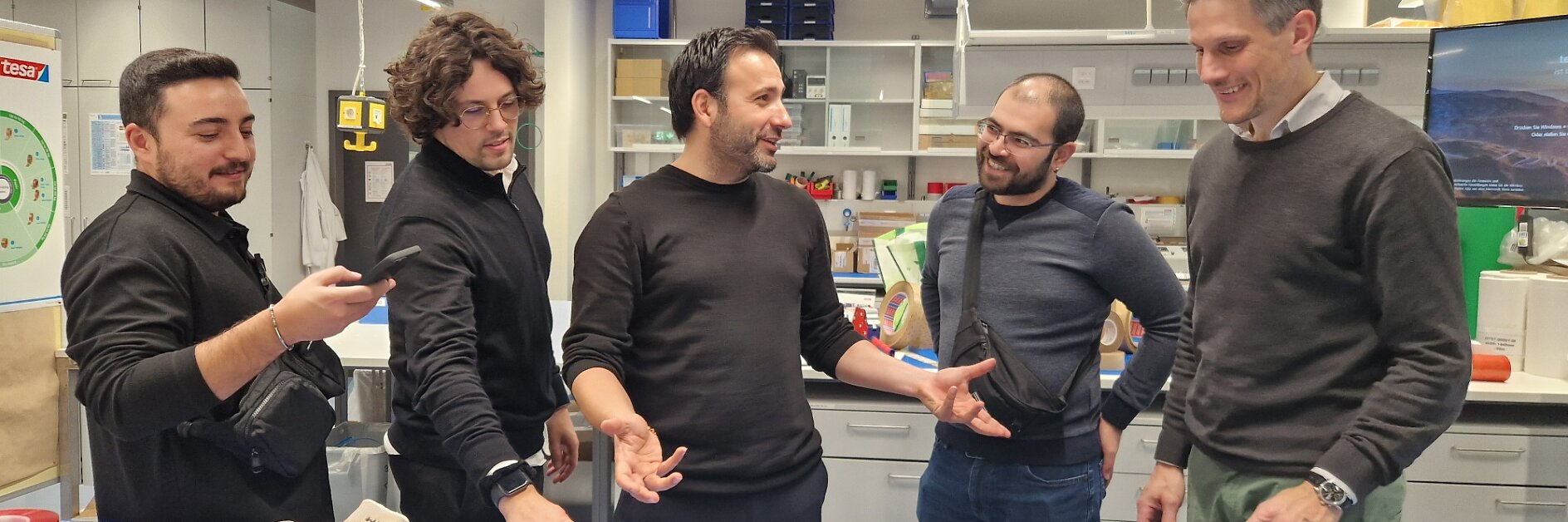 Five men stand around a worktable in a modern lab or workshop, engaging in conversation. One man checks his phone, and various equipment, gloves, tools, and rolls of tesa tape are visible on the table. Shelves and monitors are in the background. (This text has been generated by AI)