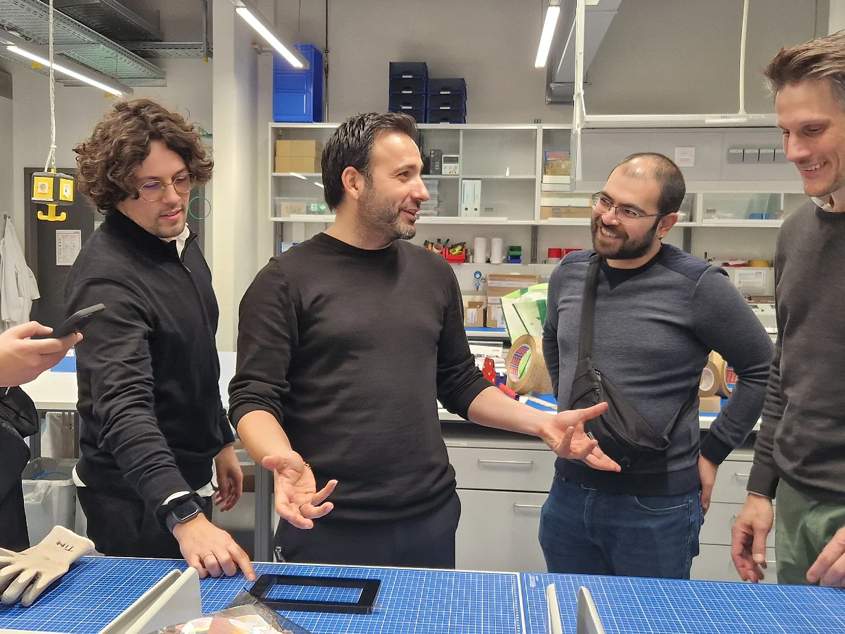 Five men stand around a worktable in a modern lab or workshop, engaging in conversation. One man checks his phone, and various equipment, gloves, tools, and rolls of tesa tape are visible on the table. Shelves and monitors are in the background. (This text has been generated by AI)