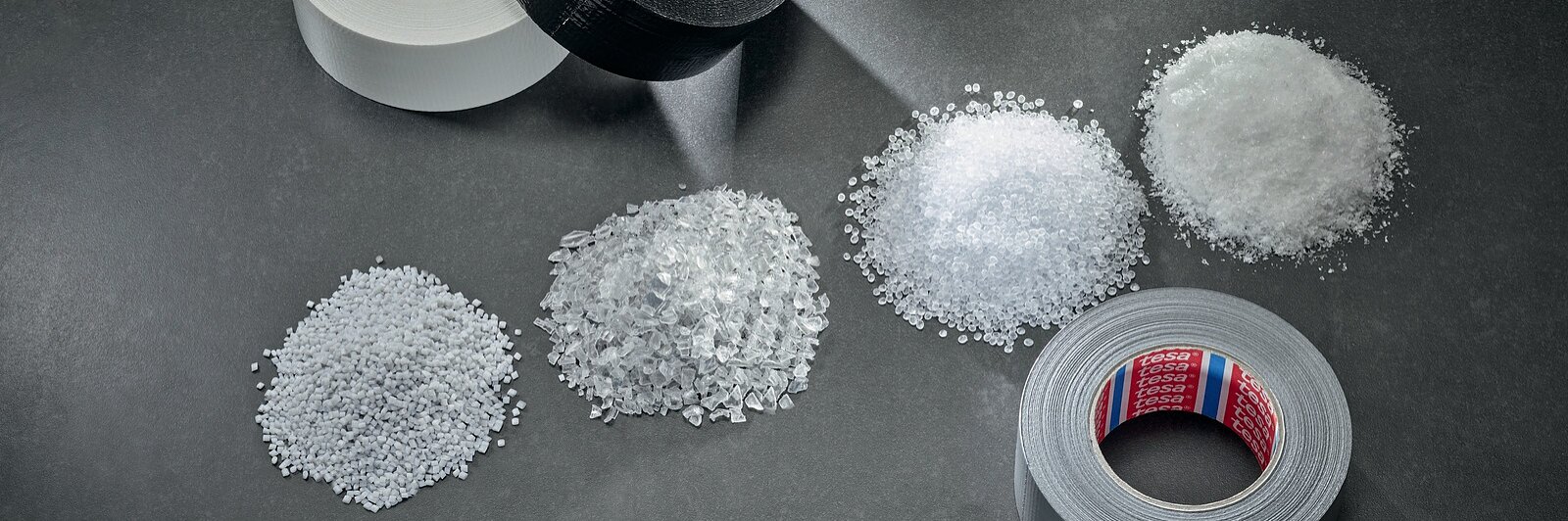 Three rolls of tesa tape—white, black, and gray—are arranged on a gray surface. In front of them are five separate piles of clear and white plastic granules and shards, displayed in varying textures and sizes. (This text has been generated by AI)