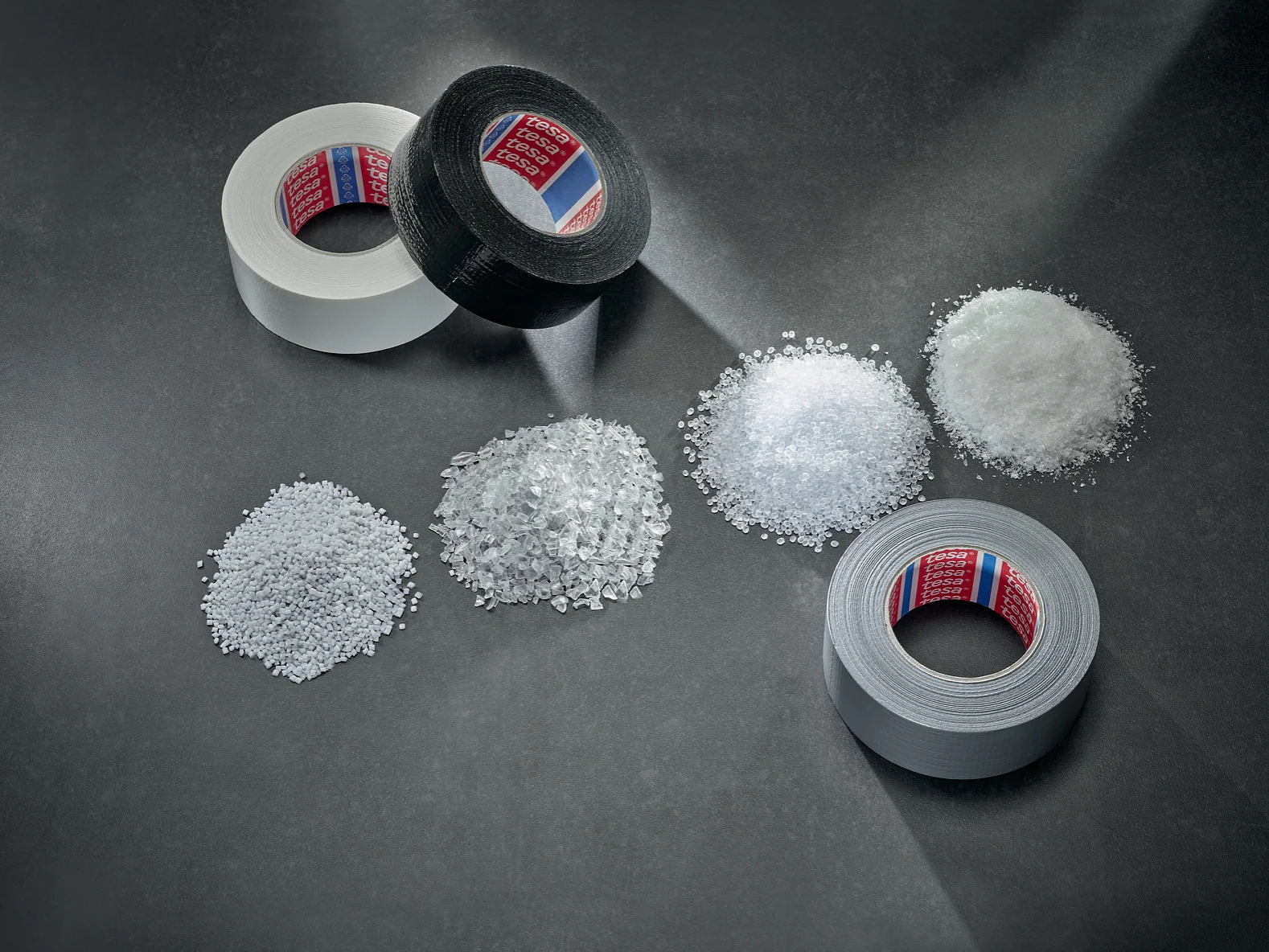 Three rolls of tesa tape—white, black, and gray—are arranged on a gray surface. In front of them are five separate piles of clear and white plastic granules and shards, displayed in varying textures and sizes. (This text has been generated by AI)