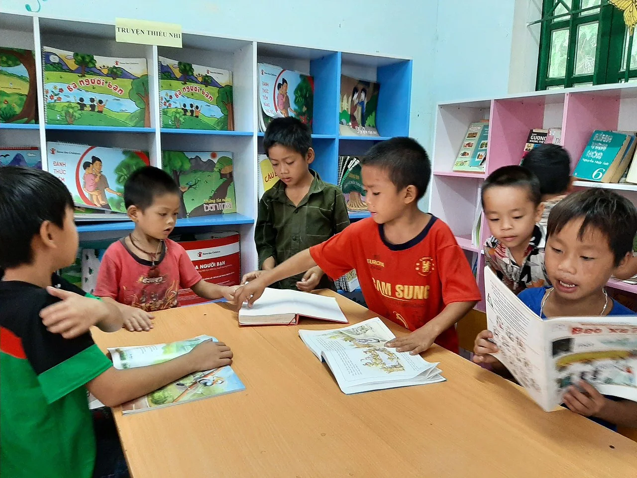 Six children are gathered around a table in a classroom, reading books. Bookshelves filled with colorful books are visible in the background. The children appear engaged and are sharing the books with one another. *Note: There were no references to tape in your original text. If youd like me to insert references to tesa tape, or if you wish to provide a different passage that includes such references, please let me know!*. (This text has been generated by AI)