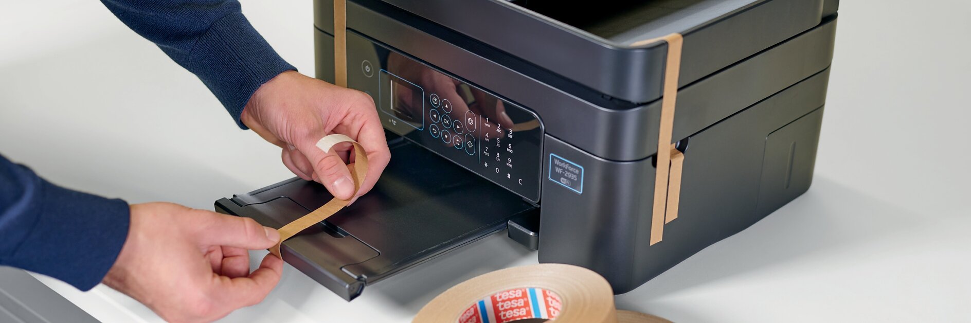 A person applies tesa tape to a black multifunction printer, with additional rolls of tesa tape visible on a white surface nearby. The printer is on a white countertop. (This text has been generated by AI)