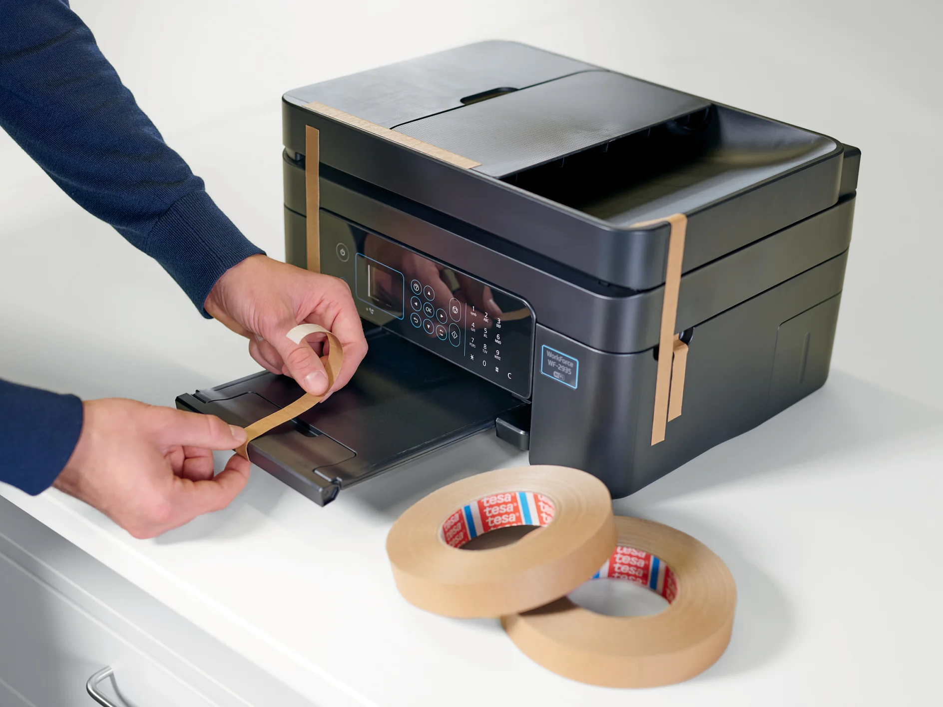 A person applies tesa tape to a black multifunction printer, with additional rolls of tesa tape visible on a white surface nearby. The printer is on a white countertop. (This text has been generated by AI)