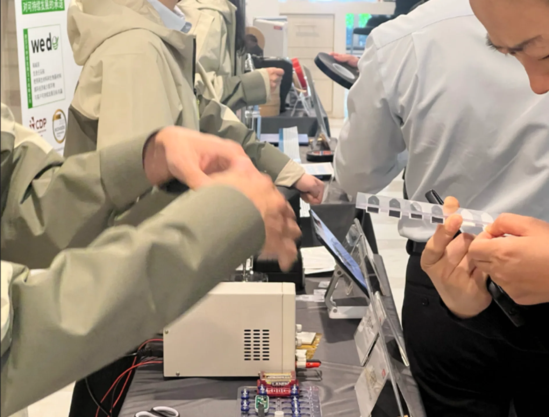 People stand at a table with electronic devices, wires, and display cards. One person examines a transparent object secured with tesa tape while others interact and gesture near the equipment. (This text has been generated by AI)