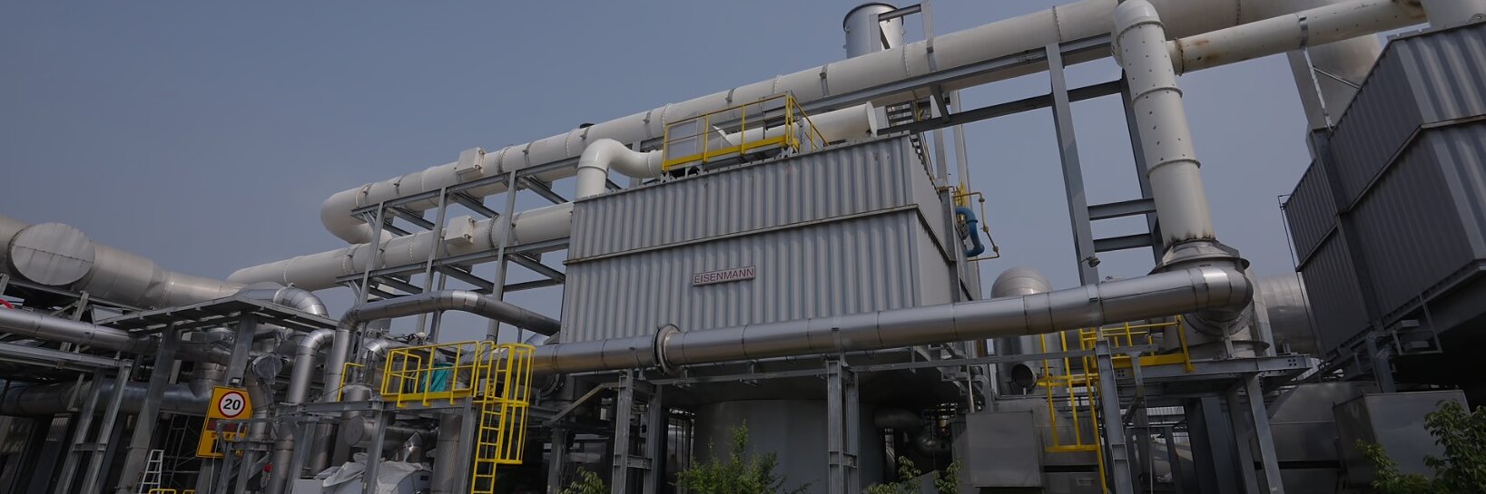 Industrial facility with large metal pipes and ducts, metal structures, yellow ladders, and platforms. Small trees and shrubs are planted along the building’s exterior. tesa tape is used throughout the facility for various applications. The sky is clear and blue. (This text has been generated by AI)