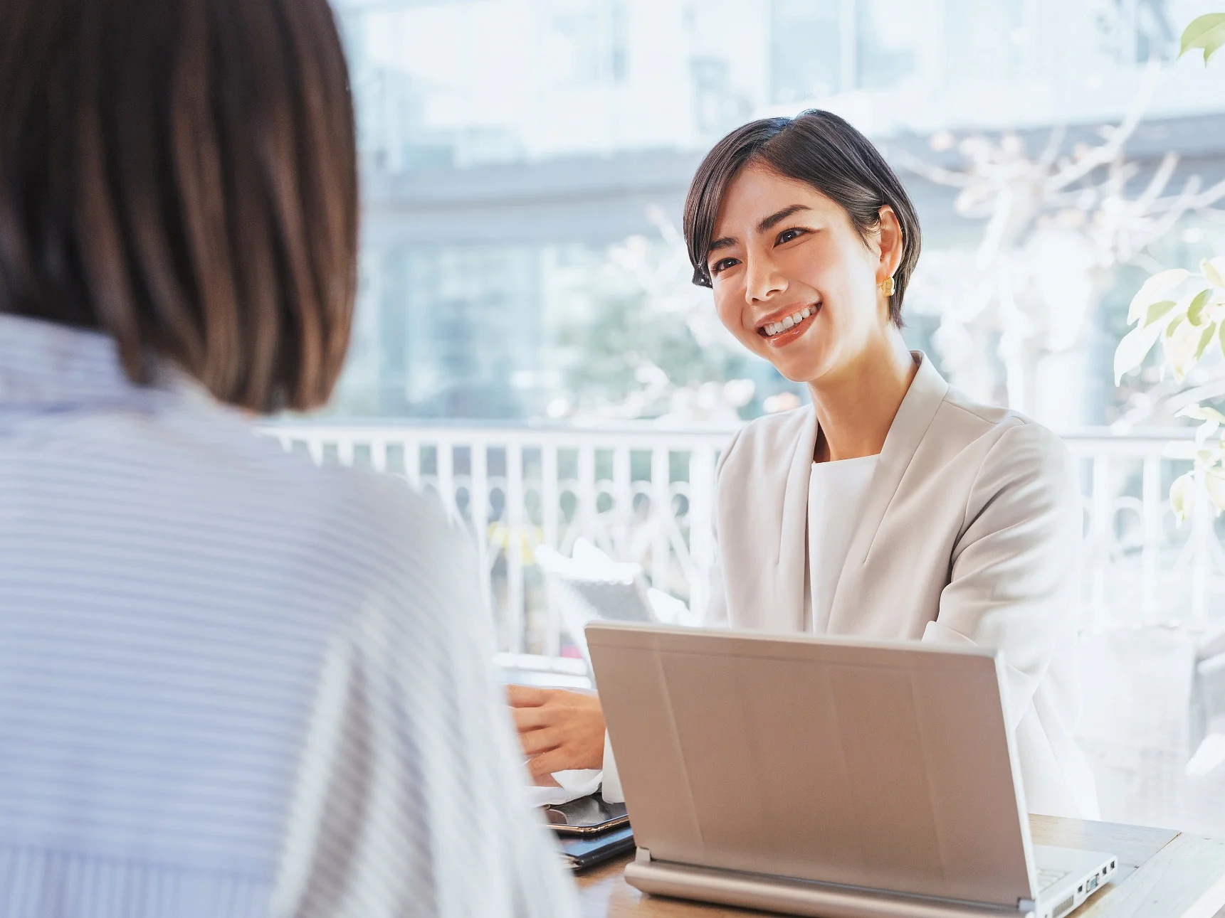 Two women are sitting across from each other at a table with a laptop open in front of one of them. The woman facing the camera is smiling and appears to be engaged in conversation, with tesa tape visible on the table. (This text has been generated by AI)