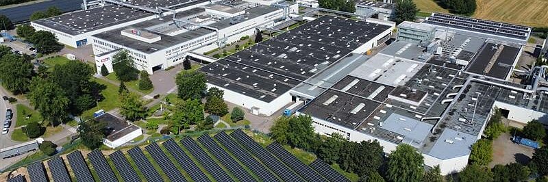 Aerial view of an industrial complex with large white buildings, surrounded by trees and fields, featuring a tesa tape solar panel array installed on the ground in the foreground. (This text has been generated by AI)