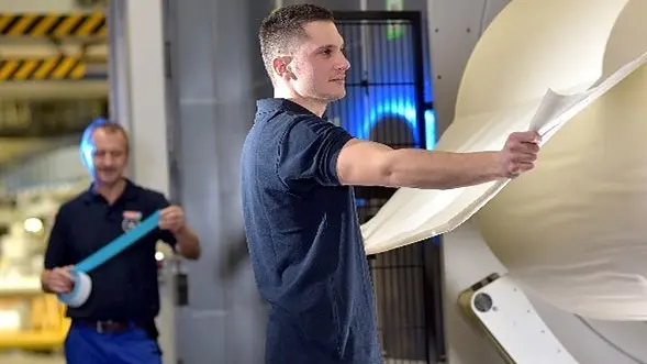 Two workers in a factory handle large rolls of white material. One man in the foreground inspects the sheet, while another in the background holds a roll of blue tesa tape. Both wear dark shirts and blue pants. Industrial equipment is visible.