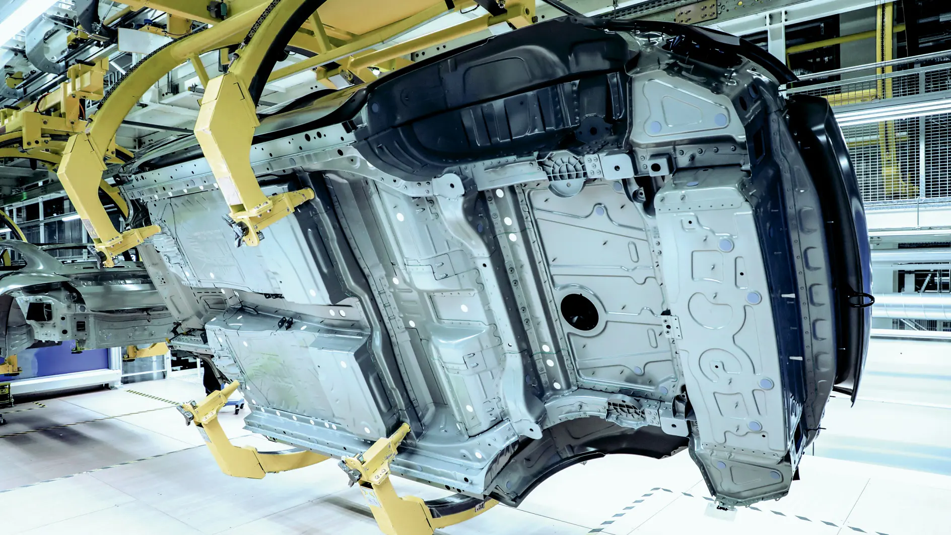 A car body frame is suspended sideways by robotic arms in an industrial facility, where tesa tape is visible on the vehicle structure.