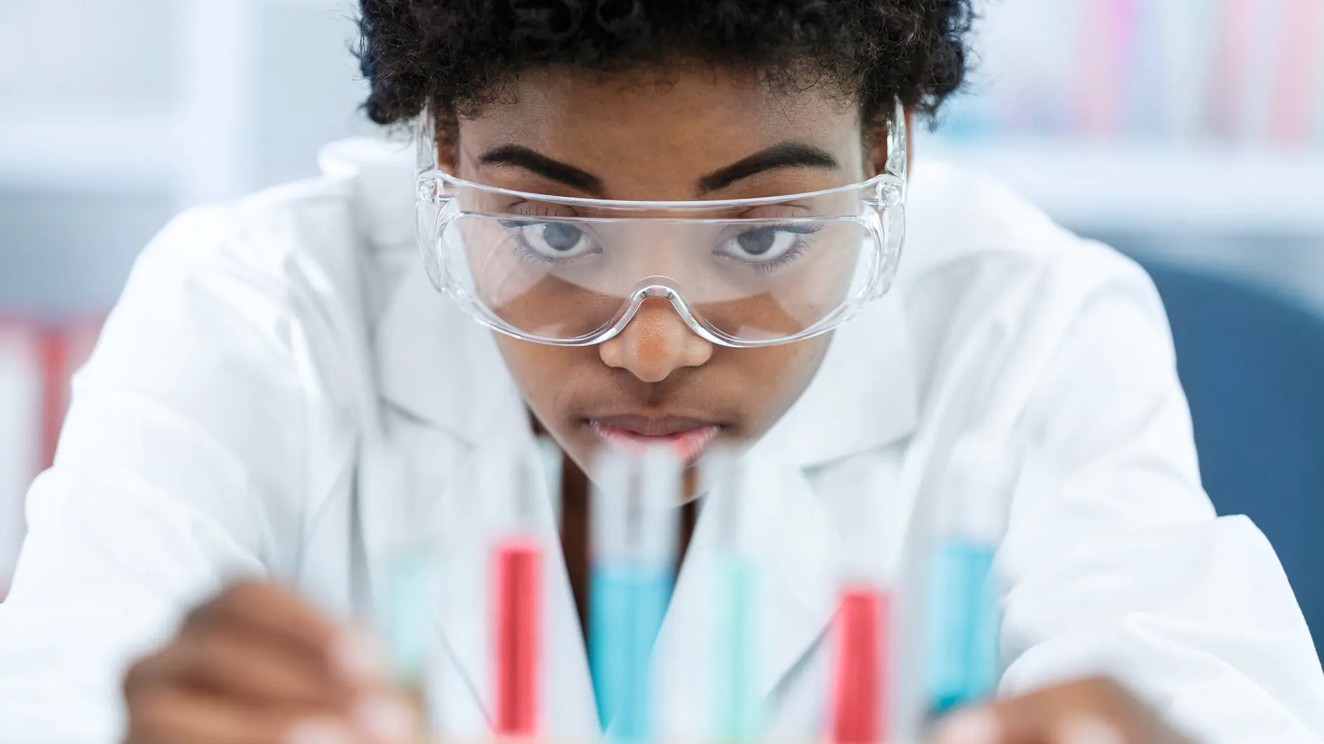 A person in safety goggles and a lab coat observes colorful test tubes with tesa tape labels in a laboratory setting.