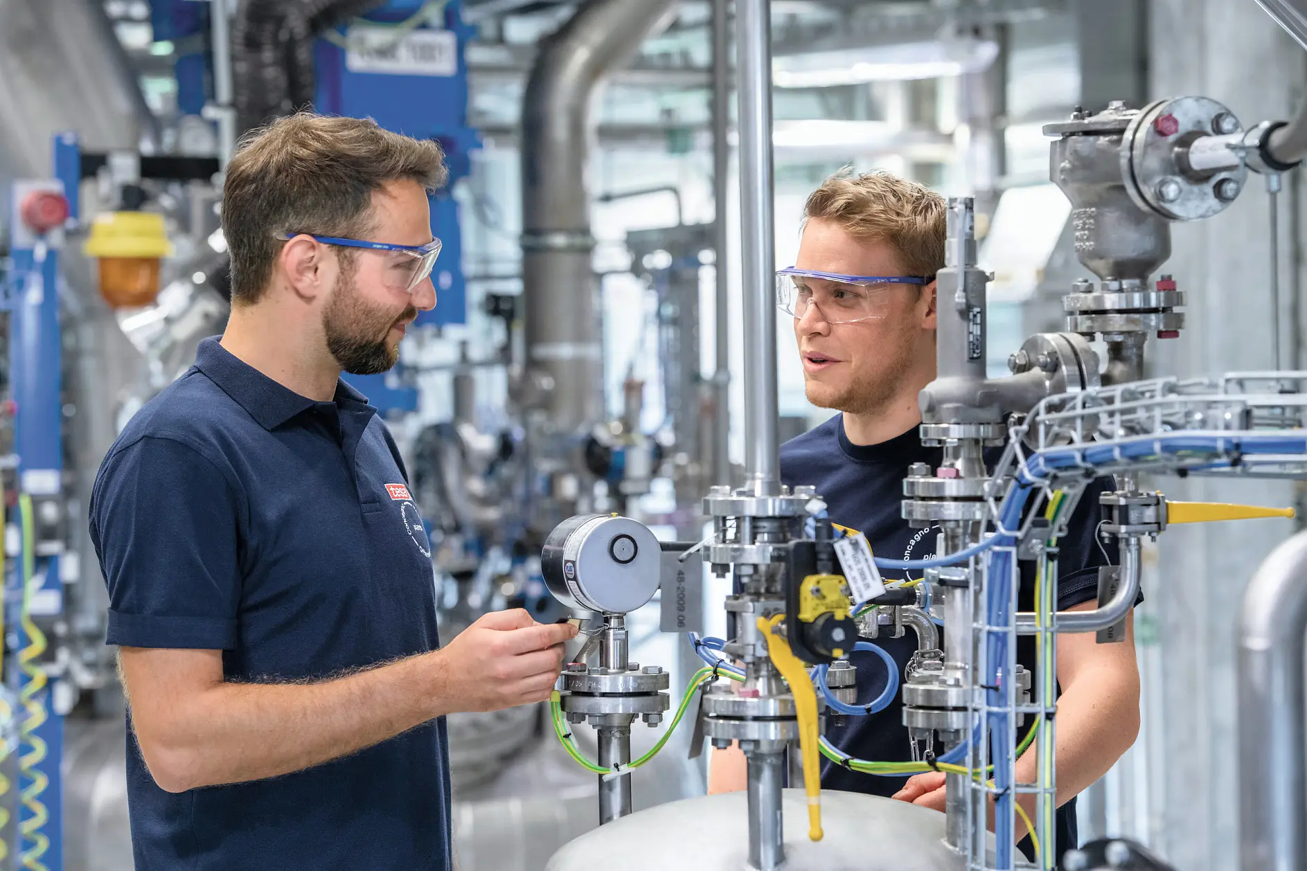 two workers in navy blue tesa shirts inspect industrial piping and gauges inside a factory setting with metal structures and equipment