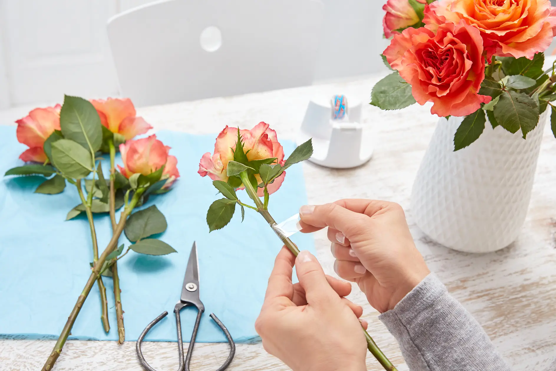 Hands are seen preparing roses by wrapping tesa tape around their stems. Several roses and a pair of scissors are on a table with a blue cloth. A vase with red and orange roses is in the background.