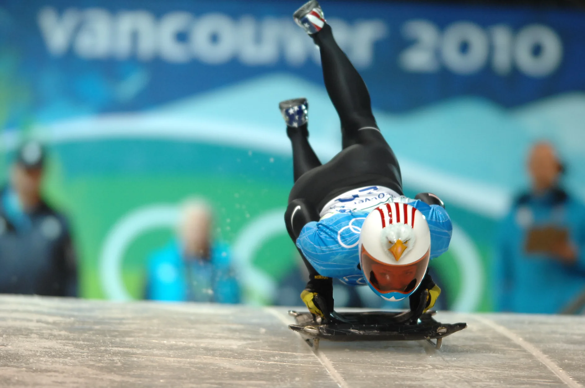 DSC_3556.JPG An athlete in a blue and white suit and red and white helmet competes in a skeleton event on an ice track. The event occurs during the Vancouver 2010 Winter Olympics, as indicated by the blurred signage in the background. (This text has been generated by AI)