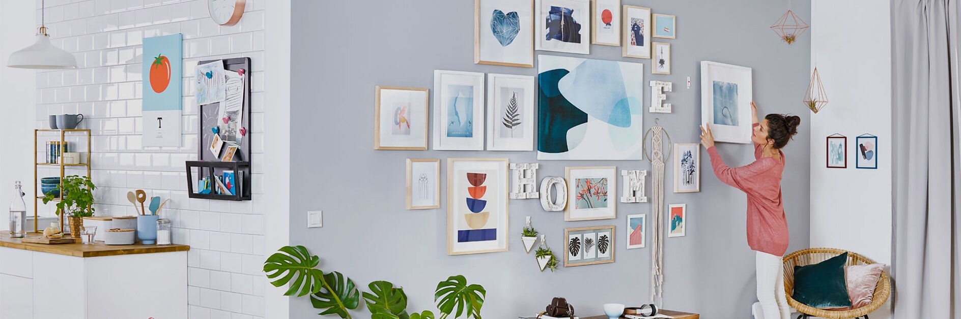 A woman in a pink shirt is arranging frames on a grey and white wall, filled with various artwork and photos, using tesa tape. The room has a modern design with a small kitchen on the left, light wood flooring, and green plants for decoration. (This text has been generated by AI)