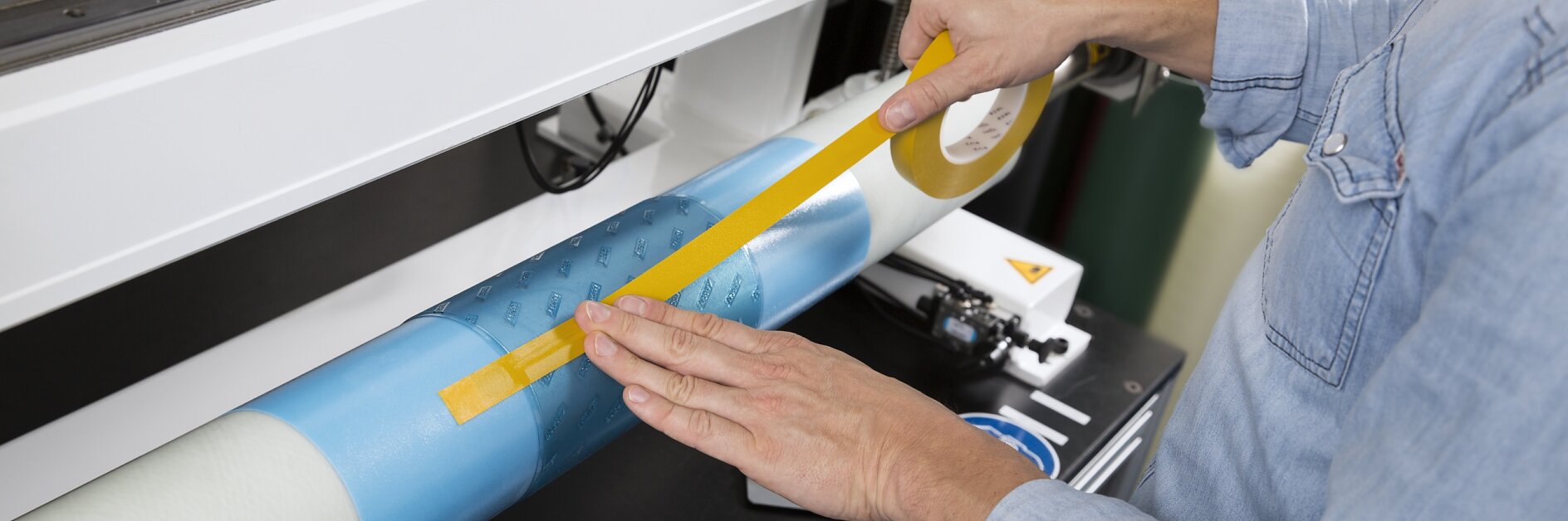 A person is wrapping a cylindrical object with blue and clear film using yellow tesa tape. The person is wearing a denim shirt and is standing at a workstation with various tools and equipment visible in the background. (This text has been generated by AI)