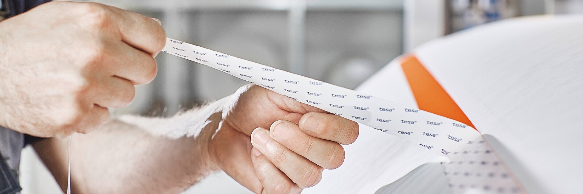 Close-up of a persons hands holding and inspecting a strip of tesa tape. The background includes industrial equipment and shelves, slightly out of focus. (This text has been generated by AI)