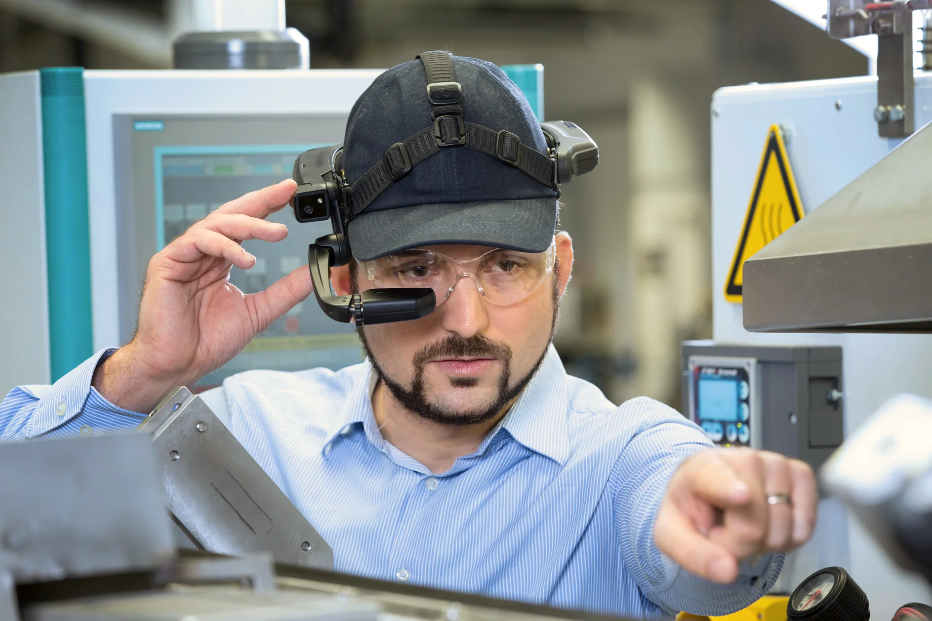 A man standing in an industrial setting wears a dark cap, safety glasses, and a headset with a visor. He appears to be focused, gesturing with his left hand and touching the visor with his right. Machinery and a screen are visible in the background. Note that any mention of tape has been replaced with tesa tape. (This text has been generated by AI)