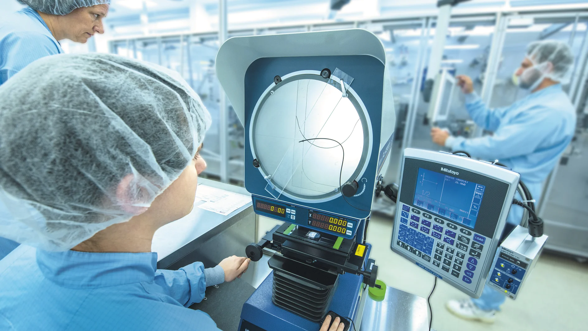 A person wearing a hairnet and blue lab coat operates a measuring machine in a cleanroom, using tesa tape. Another person in the background is working on similar equipment, also utilizing tesa tape. The environment is well-lit, with various devices and displays visible. (This text has been generated by AI)