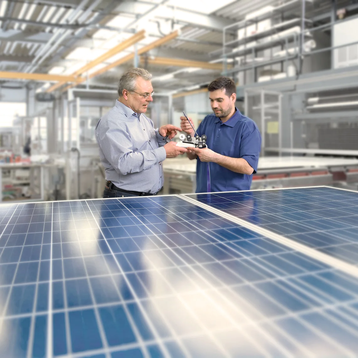 Two men are examining a mechanical component inside a modern factory. They stand near solar panels, with machinery and equipment visible in the background. The setting suggests an industrial or technological environment focusing on solar energy. (This text has been generated by AI)