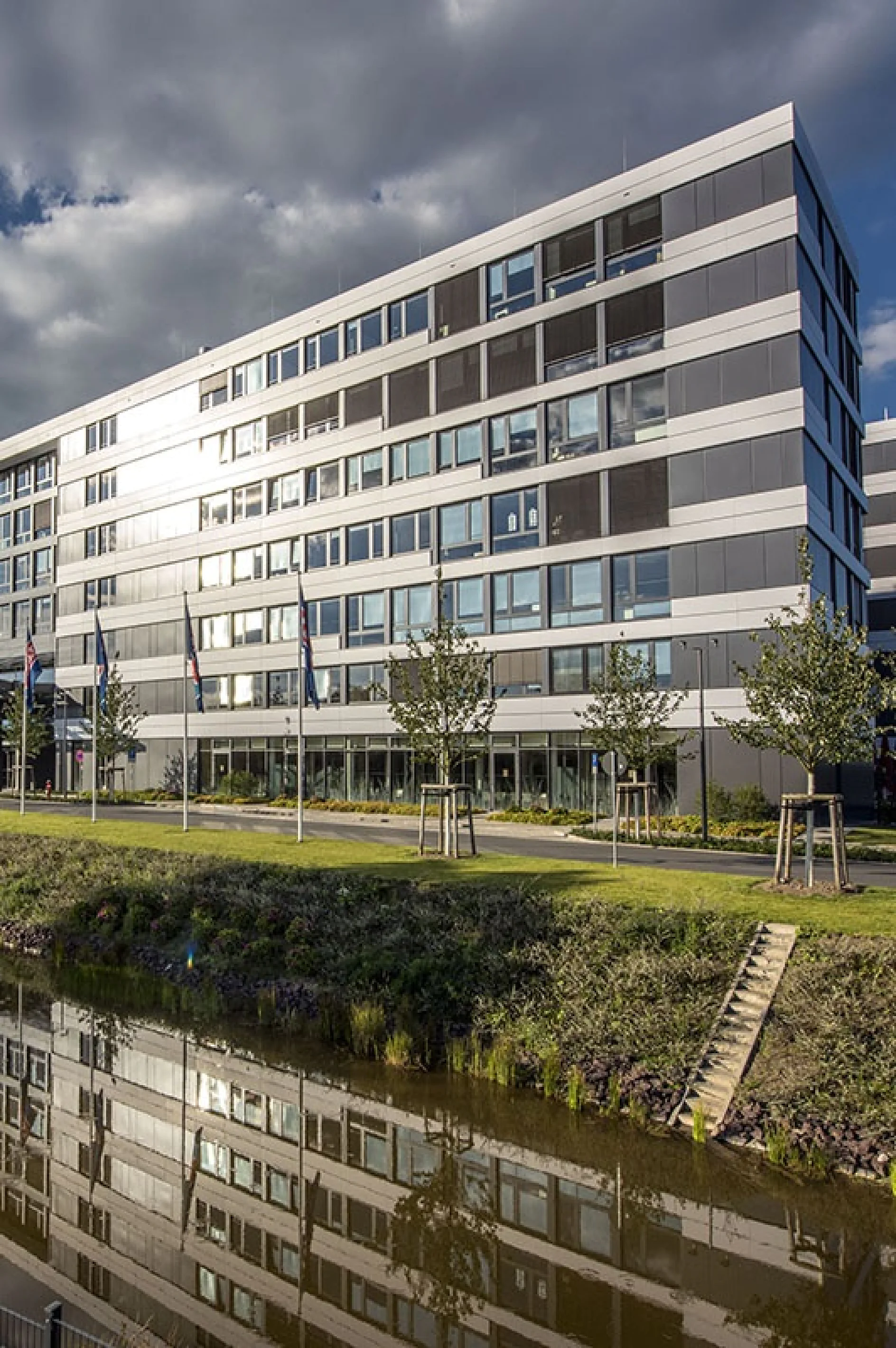 A modern, multi-story office building with a sleek facade and large windows is reflected in a calm body of water. Flags line the front, and trees are planted along a grassy area. The sky is partly cloudy. (This text has been generated by AI)