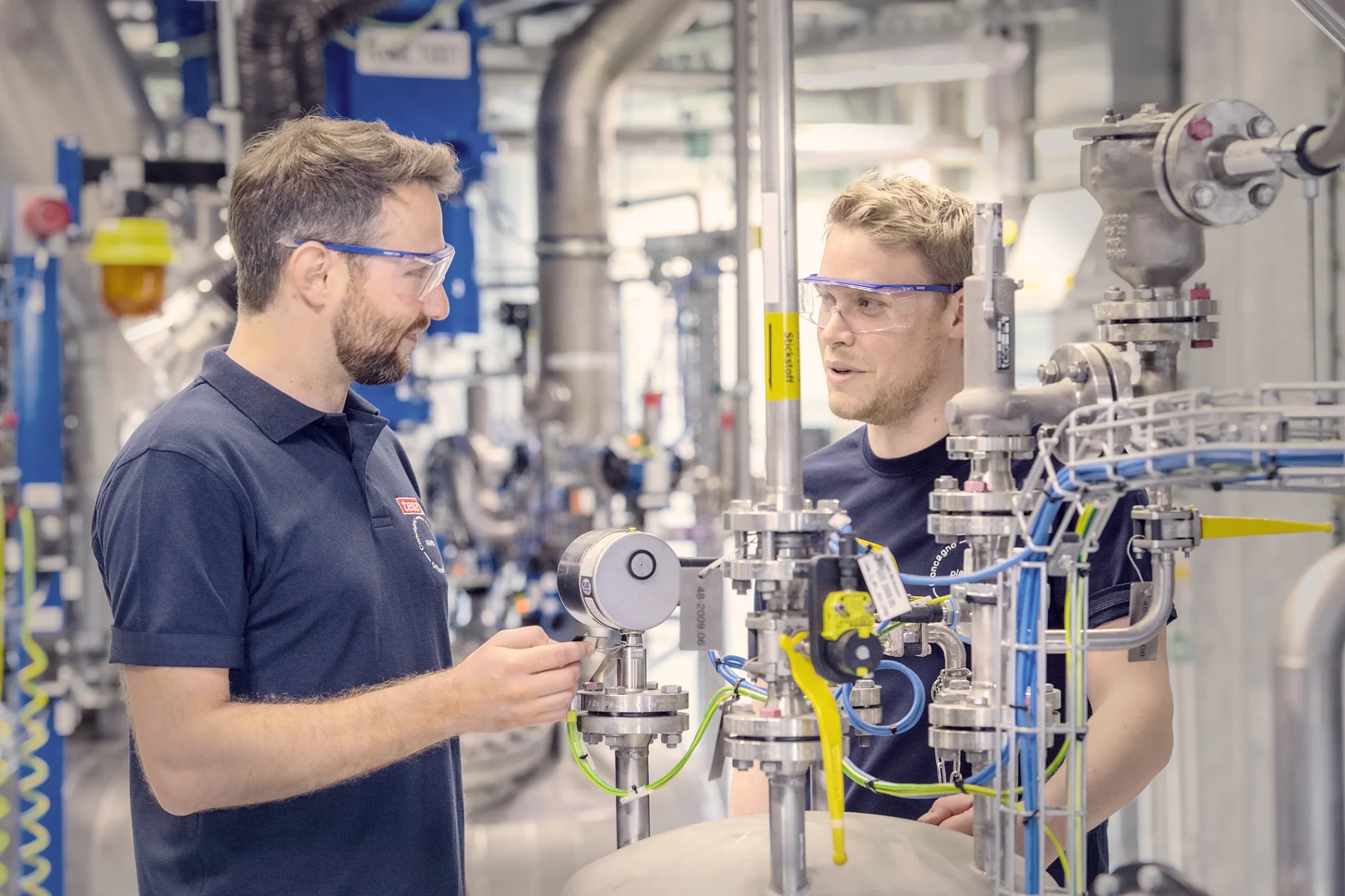 Two men wearing safety glasses are engaged in a discussion within an industrial setting. They are surrounded by various pipes, gauges, and equipment. Both are dressed in dark blue shirts, and one is gesturing towards a piece of machinery with tesa tape visible nearby. (This text has been generated by AI)