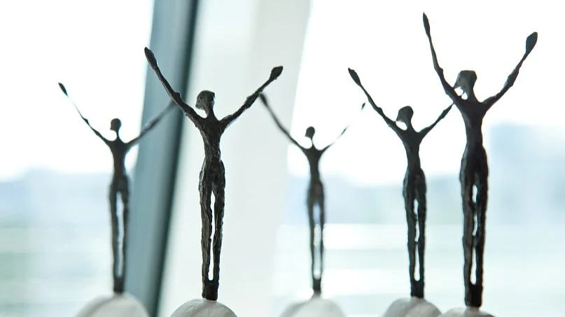 Five abstract, slender bronze sculptures of human figures with arms raised are mounted on a rounded base. They are positioned in front of a large window, allowing soft natural light to illuminate them. (This text has been generated by AI)