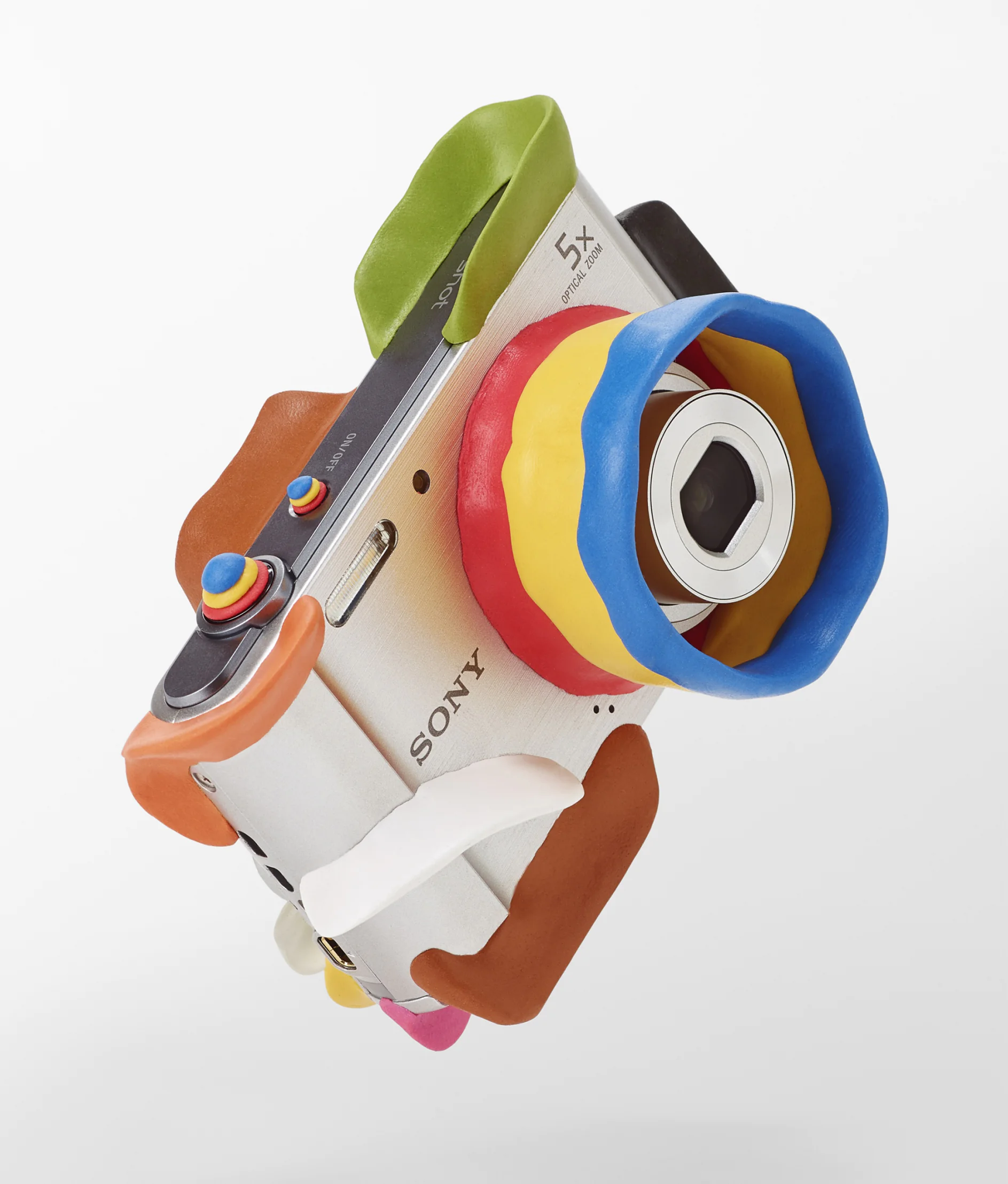 A digital camera wrapped in colorful playdough, featuring multiple bright colors including blue, red, yellow, and green. The camera lens and controls are partially covered with tesa tape, adding a playful appearance. The background is plain white. (This text has been generated by AI)