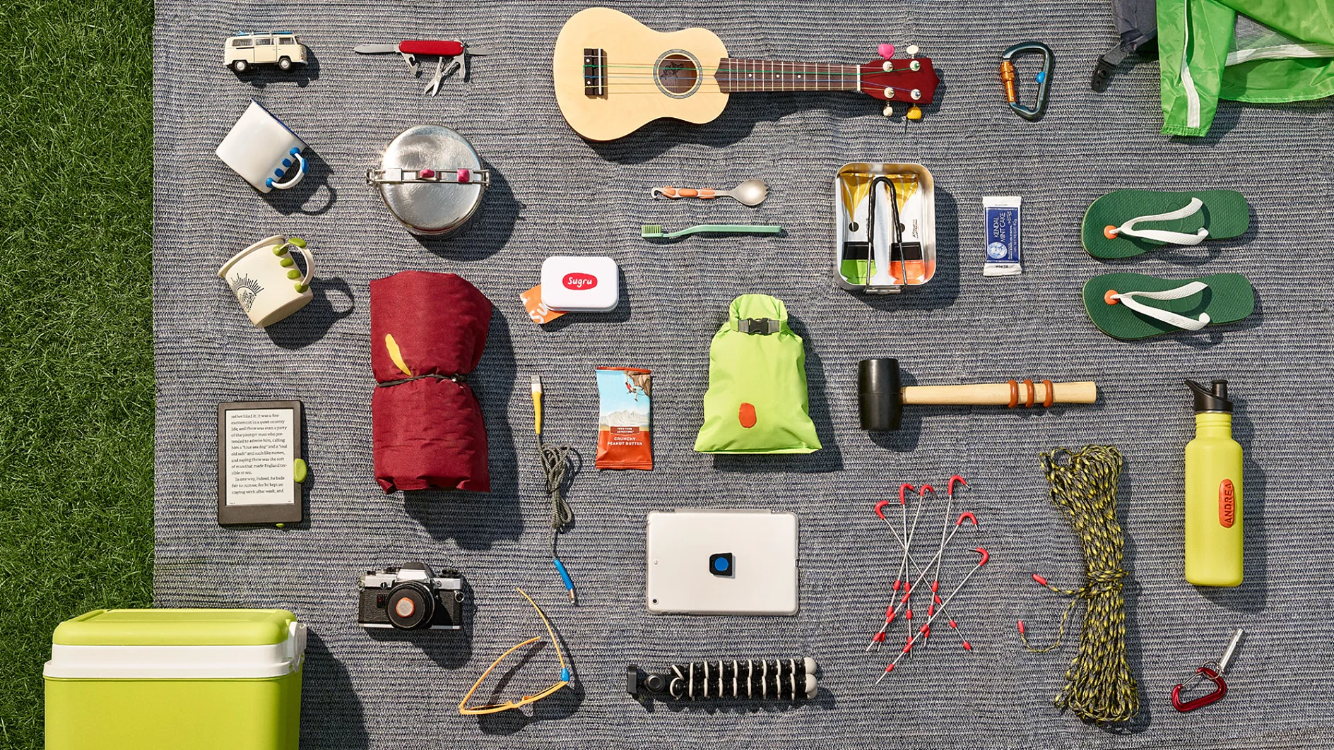 A neatly arranged collection of camping items on a gray blanket: a ukulele, hammock, carabiners, flip-flops, cooler, kettle, sunscreen, books, camera, tablet with tesa tape attached for protection and support during an outdoor adventure. Additionally included are a flashlight secured with tesa tape for durability in rugged conditions; ropes reinforced with tesa tape to ensure reliability; and a water bottle. Grass borders the blanket on the left side. (This text has been generated by AI)